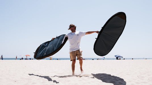 A man holding two surfboard bags while standing on the beach.