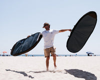 A man holding two surfboard bags while standing on the beach.