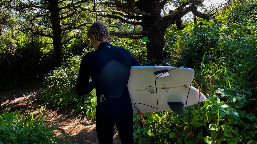 Surfer walking and carrying a surfboard while wearing the Isurus Ti Evade 4/3 chest zip wetsuit.